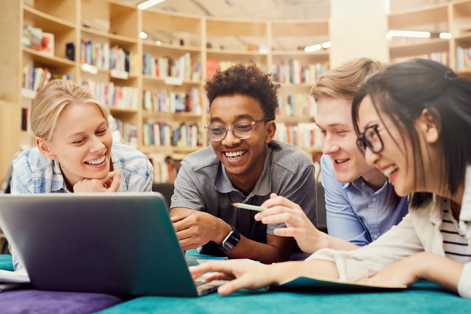 Positive excited multi-ethnic students in casual clothing lying on floor in campus library and laughing while watching curious video on laptop